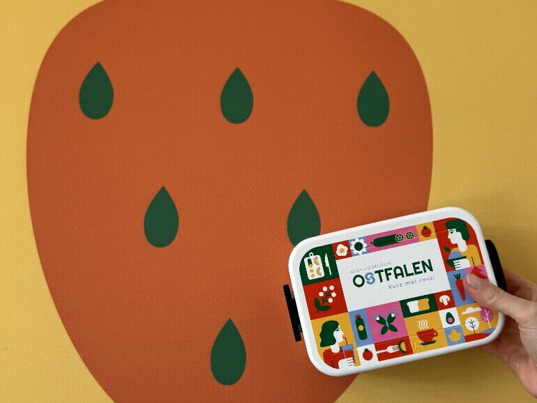 Eine Hand hält eine bunte Brotdose mit der Aufschrift OSTFALEN Kurz mal raus vor einer großen stilisierten