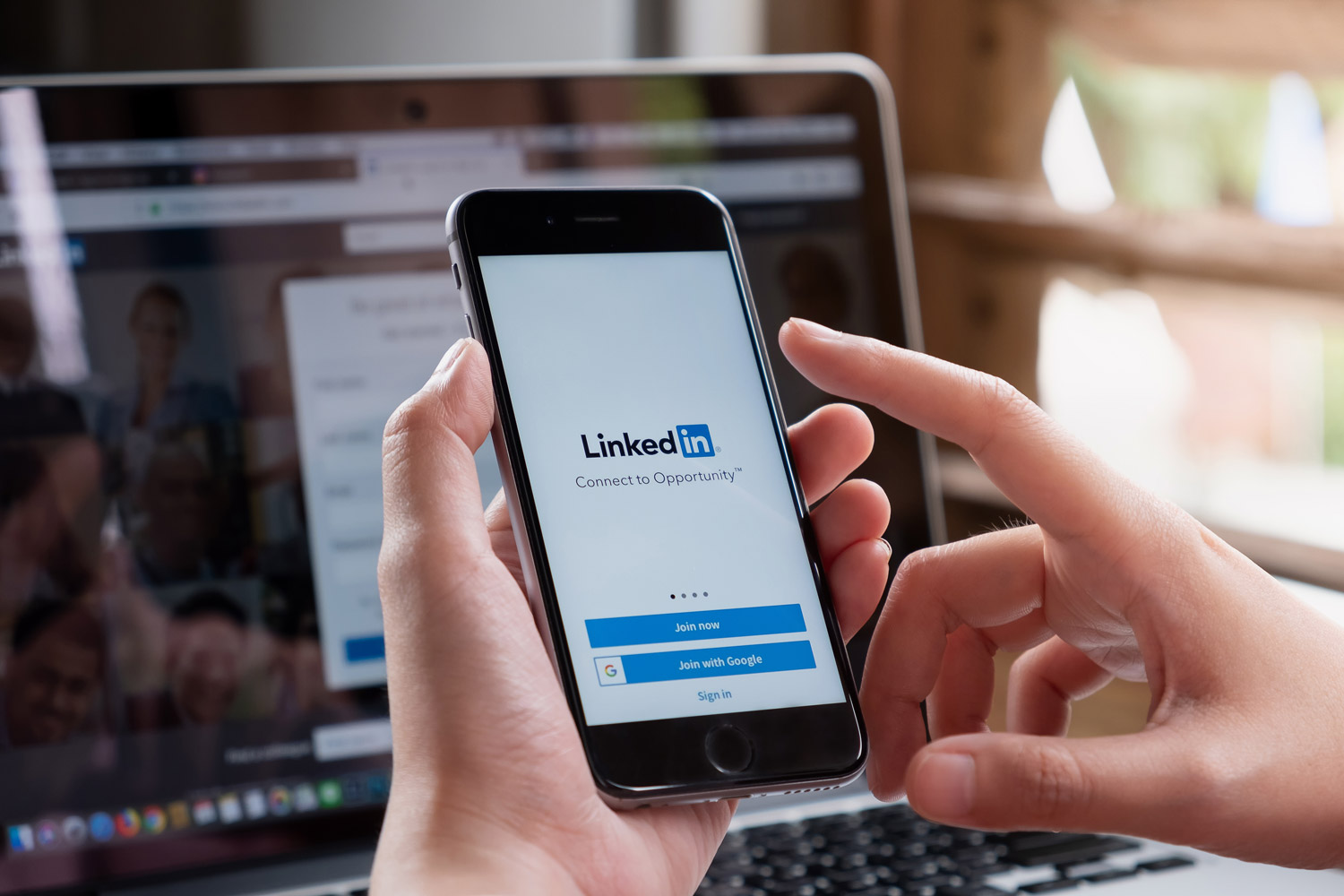 Eine Person hält ein Smartphone mit der LinkedIn-Anmeldeseite, im Hintergrund ist ein Laptop zu sehen