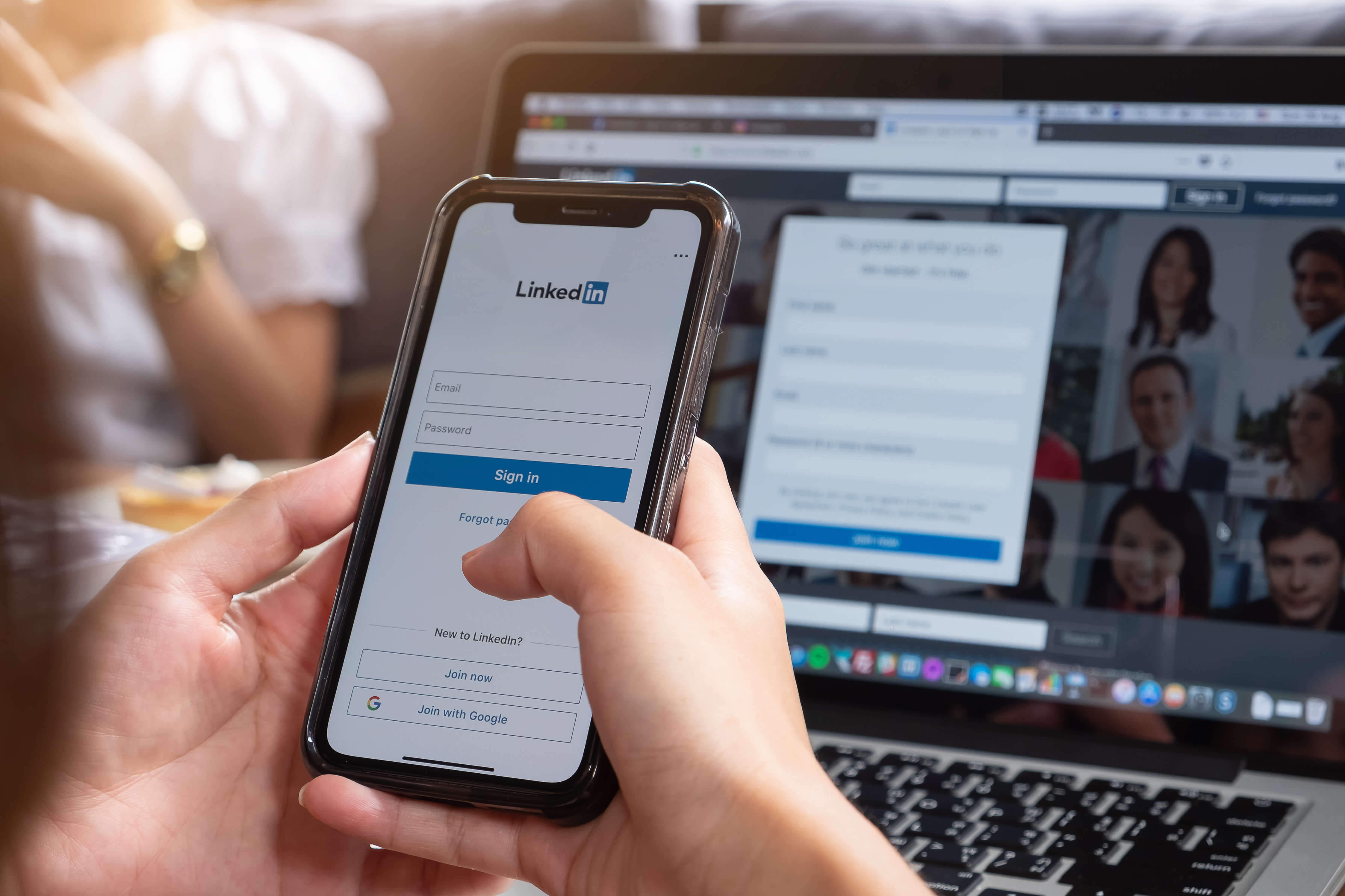 Eine Person benutzt ein Smartphone mit der LinkedIn-Anmeldeseite, während im Hintergrund ein Laptop mit der